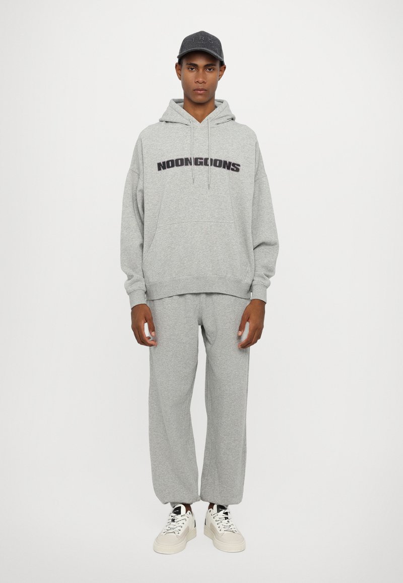 Felpa grigia con testo "NOONGOONS", abbinata a pantaloni della tuta grigi coordinati. Indossata con sneakers bianche, dotate di lacci rotondi e un cappellino da baseball.