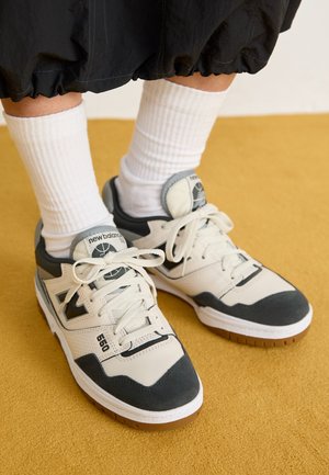 Osoba nosząca białe i szare sneakersy New Balance 550 z białymi skarpetami za kostkę, stojąca na musztardowozółtym dywanie.