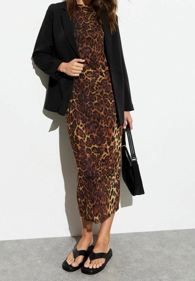 Femme portant une longue robe imprimé léopard, un blazer noir, des sandales noires, et portant un sac à main noir, se tenant contre un mur blanc.