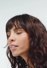 Kad su uži plan, osoba s dugom, tamnom, kovrčavom kosom i svijetlom kožom, s glatkom kožom i prirodnim makeupom, gleda malo sa strane.