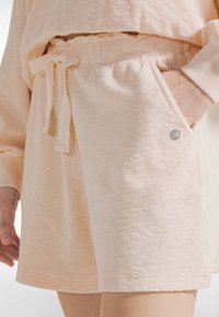 Pantalones cortos de color durazno claro hechos de tela suave tipo toalla, con una cintura elástica con cordón y bolsillos laterales. Detalle de pequeño logo presente.