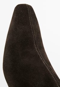 Chaussure en daim marron à bout pointu avec des coutures contrastantes et une texture lisse, mettant en avant un design épuré et minimaliste.