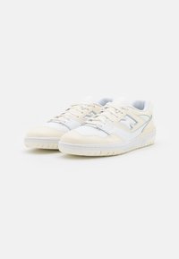 Un paio di sneaker basse bianche e beige con lacci, pannelli laterali perforati e un logo sul lato, posizionate su uno sfondo bianco.