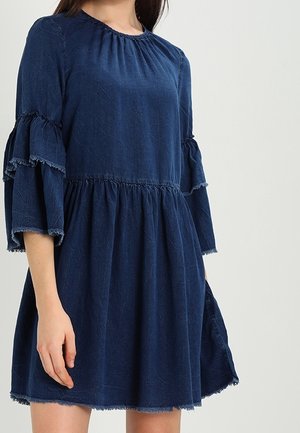 Woman wearing a dark blue, loose-fitting dress with tiered, ruffled three-quarter sleeves and a gathered waist.