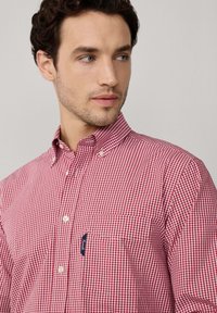 Homme à la peau claire et aux cheveux bouclés foncés portant une chemise à carreaux rouge et blanc avec une poche poitrine, regardant légèrement sur le côté.