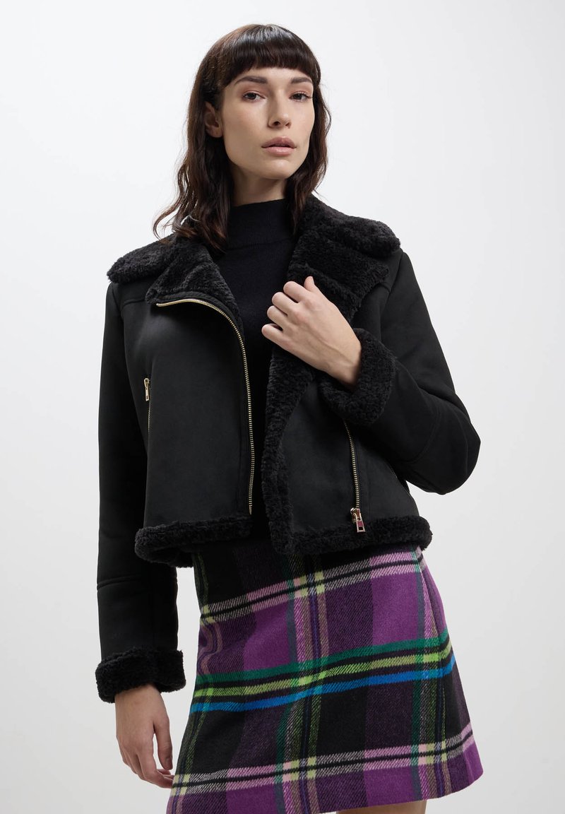 Giacca in finta shearling nera con un ampio colletto e chiusura a zip; presenta una fodera morbida e un design cropped. Abbinate a una gonna a quadri.