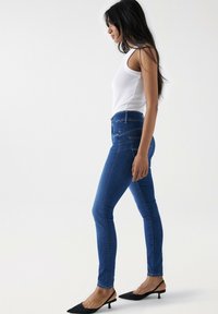 Vaqueros ajustados azules de cintura alta, con sutiles detalles de costura y bolsillos traseros, combinados con una camiseta sin mangas blanca ajustada y mules negros.