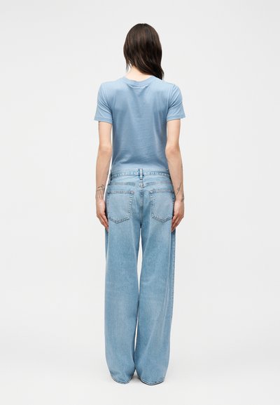 Άτομο με μακριά σκουρόχρωμα μαλλιά που φοράει γαλάζιο κοντομάνικο πουκάμισο και φαρδιά γαλάζια τζιν, στέκεται γυρισμένο προς τα πίσω σε απλό φόντο.