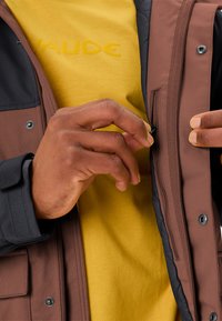 Giacca isolante marrone e nera con una tasca con zip, abbinata a una camicia gialla con logo in rilievo. Primo piano di una mano che regola la giacca.