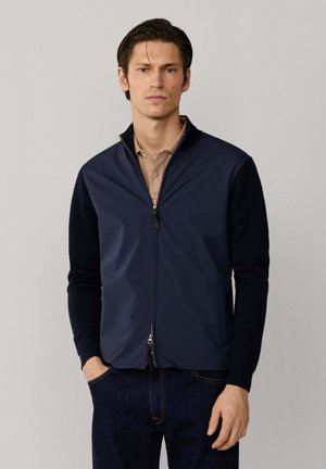 Hombre con chaqueta azul marino con cremallera y mangas negras sobre polo beige, de pie con las manos en los bolsillos frente a un fondo liso.