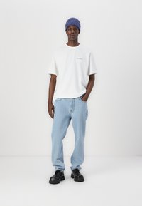 Tricou alb cu mânecă scurtă, cu text imprimat, blugi cu picior drept de culoare albastru deschis și pantofi negri cu talpa groasă, completat cu o căciulă albastră cu model în relief.