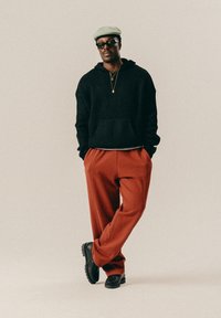 Άνδρας με μπεζ καπέλο, μαύρα γυαλιά ηλίου, μαύρο πουλόβερ, παντελόνι χάλκινου χρώματος, μαύρα παπούτσια, στέκεται με τα χέρια στις τσέπες και τα πόδια σταυρωμένα.