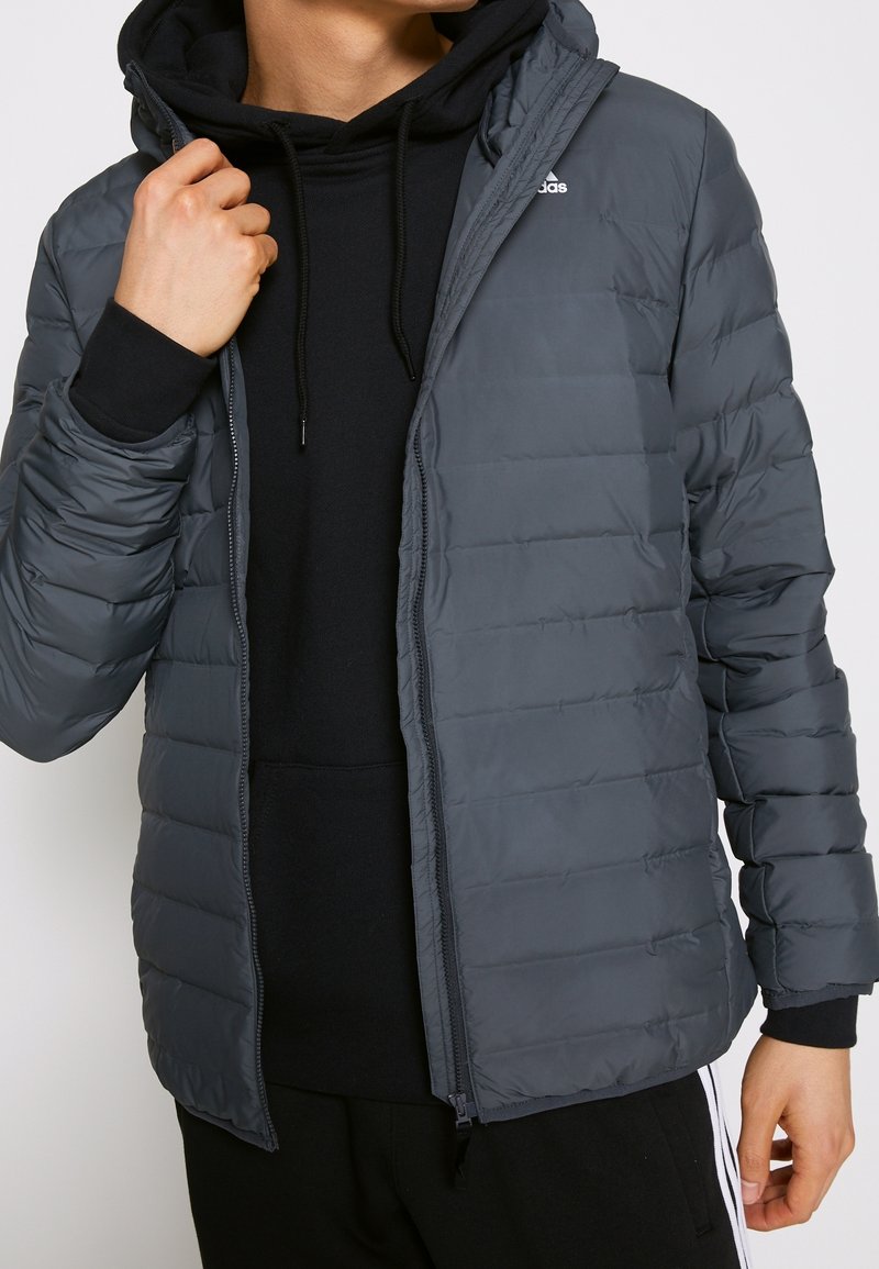 Giacca a vento grigia con zip frontale, cappuccio e texture trapuntata. Indossata sopra un felpa nera, con un logo sul petto sinistro.