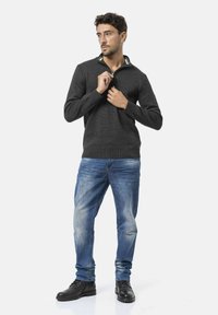 Donkergrijze trui met een ritssluiting bij de kraag, lange mouwen, gedragen over lichtblauwe jeans en zwarte schoenen. Eenvoudige, getextureerde brei stof.