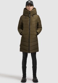 Doudoune vert olive avec un col montant, fermeture éclair à l'avant et couture horizontale. Associée à un legging noir et des bottes de combat. Casquette noire avec logo.