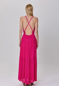 Robe longue rose en tissu léger, présentant un dos bas avec des bretelles croisées, un design à volants et une silhouette fluide.