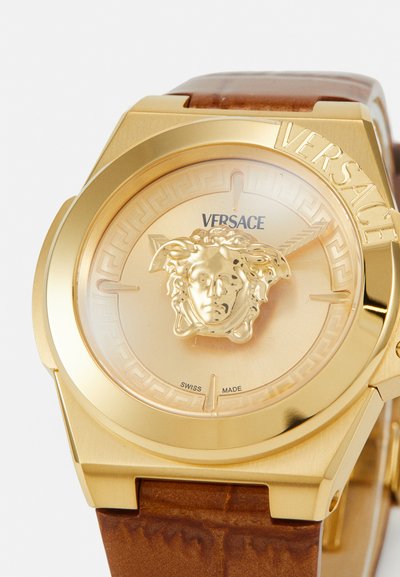Zeltīts Versace pulkstenis ar apaļu korpusu, kurā ir lauvu galva uz bēša ciparnīcas un brūna teksturēta ādas siksna.