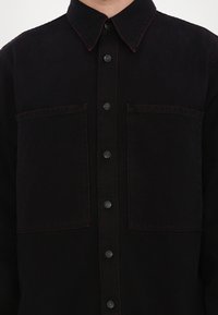 Cămașă din denim negru cu guler ascuțit, două buzunare pe piept și cusături roșii în contrast. Închidere cu nasturi negri.