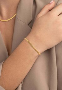 Bracciale in tonalità oro con un design sottile e texture, appoggiato su una mano, abbinato a una collana in tonalità oro coordinata su uno sfondo di tessuto beige.