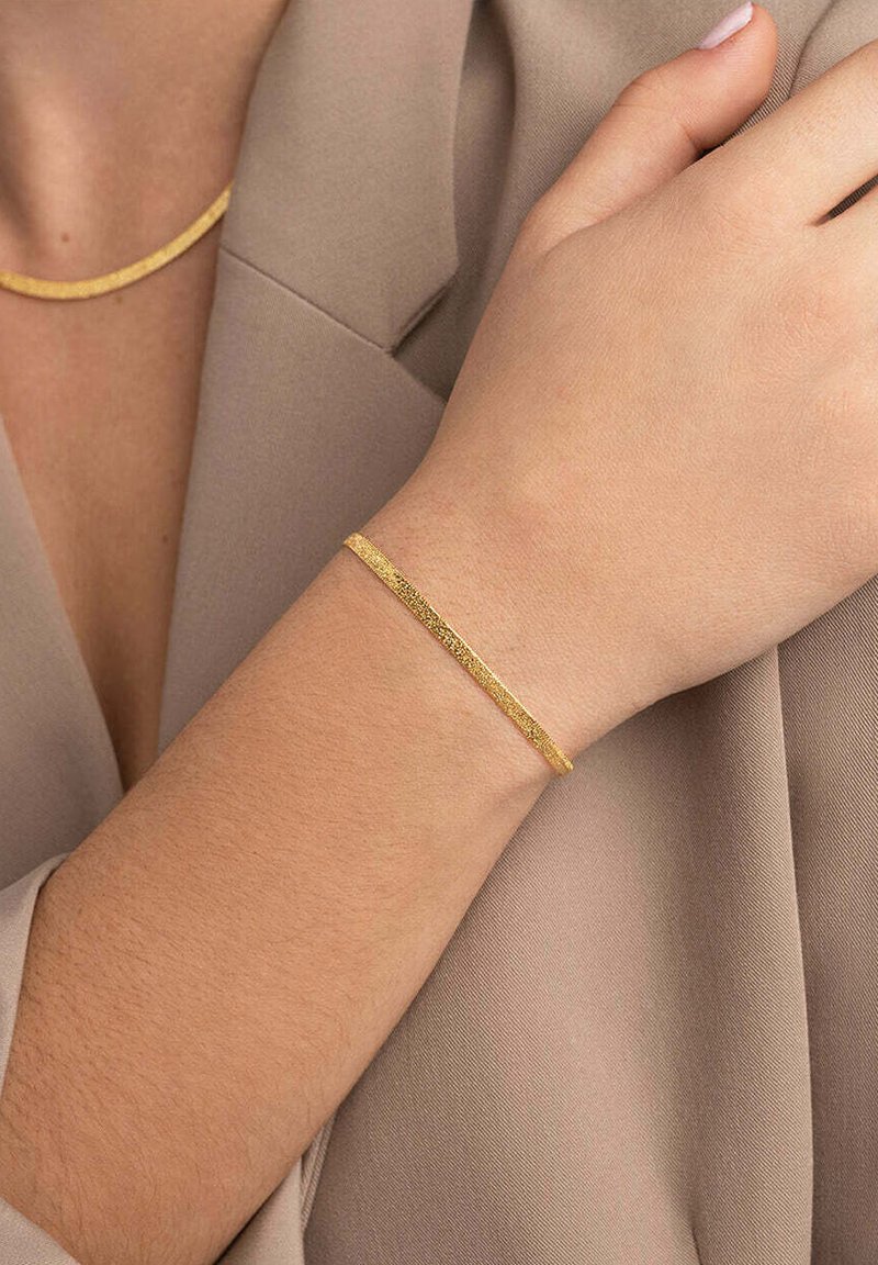Bracciale in tonalità oro con un design sottile e texture, appoggiato su una mano, abbinato a una collana in tonalità oro coordinata su uno sfondo di tessuto beige.