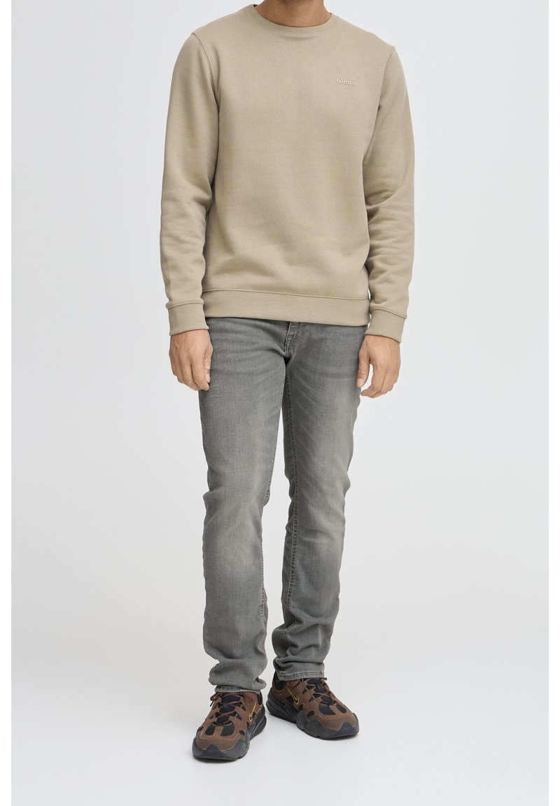 Homme portant un sweat-shirt beige à manches longues, un jean gris délavé, et des chaussures de sport marron et noires, debout devant un fond uni clair.