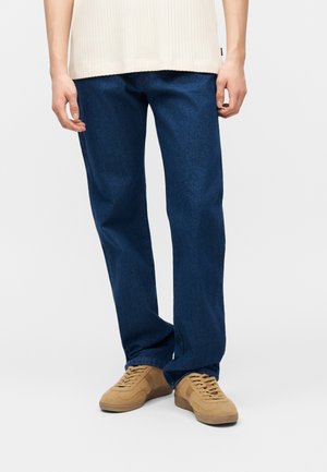 Person iført mørkeblå straight-leg jeans og beige snøresko, stående mod en ensartet lys baggrund.