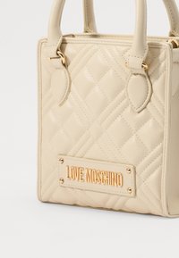 Beżowa torebka o pikowanej powierzchni z dwoma uchwytami, złotymi elementami i złotą wytłoczoną płytką z logo. Teksturowana powierzchnia z wzorem w diamenty.