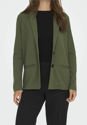 Blazer vert olive en tissu lisse, avec un col cranté, deux poches avant et une fermeture à un bouton. Porté sur un top noir.