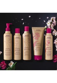Cinq produits Aveda Cherry Almond avec des flacons crème. Chaque flacon a des accents roses ; cela inclut un shampoing, un après-shampoing, une lotion pour le corps et un gel douche.