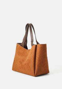 Tan suède tote tas met donkerbruin leren handvatten en witte stiksels, staande tegen een effen witte achtergrond.