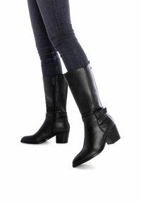 Stivali alti in pelle nera con punta affusolata, chiusura con zip laterale e una cintura decorativa intorno alla caviglia. Tacchi a blocco robusti.