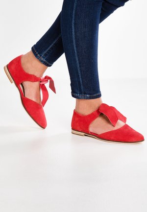 Nohy v červených semišových plochých botách s kotníkovými pásky zavázanými na mašle, doplněné tmavě modrými skinny džíny na bílém pozadí.