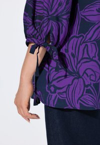 Lilla bluse med blomstermotiv på navy baggrund, med pufærmer og ties ved manchetterne. Glat stof med en afslappet pasform.