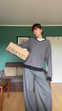Randig marinblå och vit tröja med V-ringning, tillsammans med vida grå byxor. Håller en brun Zalando-låda, placerad i ett vardagsrum.