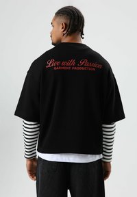 T-shirt noir oversize avec texte rouge "Vivre avec passion" et "PRODUCTION DE VÊTEMENTS" au dos. Superposé avec des manches longues rayées.