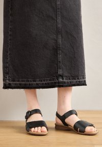 Black leather sandals with two adjustable straps, open-toe design, and a cushioned sole, styled with a denim skirt featuring a frayed hem.