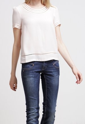 Kobieta w białej bluzce z krótkim rękawem i koronkowym wykończeniem, w dopasowanych ciemnoniebieskich dżinsach na jednolitym jasnym tle.