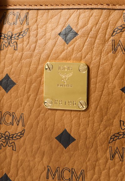 Tan leather surface with black diamond and MCM logos, centered gold metal plate engraved with "MCM München" and serial number R8196.