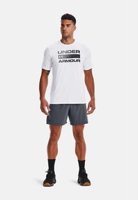 Muž stojí v bielom tričku s logom Under Armour, šedých športových šortkách, čiernych ponožkách a čiernych teniskách na bielom pozadí.