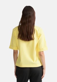 Top giallo a maniche corte con una vestibilità rilassata, realizzato in tessuto morbido. Presenta buchini arrotolati e orlo dritto, indossato sopra pantaloni neri.