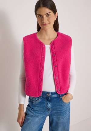 Frau trägt eine knallpinke, geknöpfte Fleeceweste über einem weißen Langarmshirt und blauen Jeans, stehend vor einem einfarbigen Hintergrund.