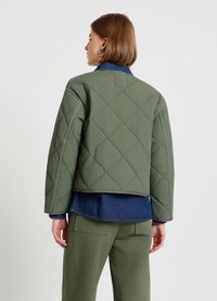 Grüne gesteppte Jacke mit kastigem Schnitt, ausgestattet mit einem blauen Denim-Kragen, sichtbaren Nähten und einem kurzen Saum, darüber getragen ein blaus Hemd.