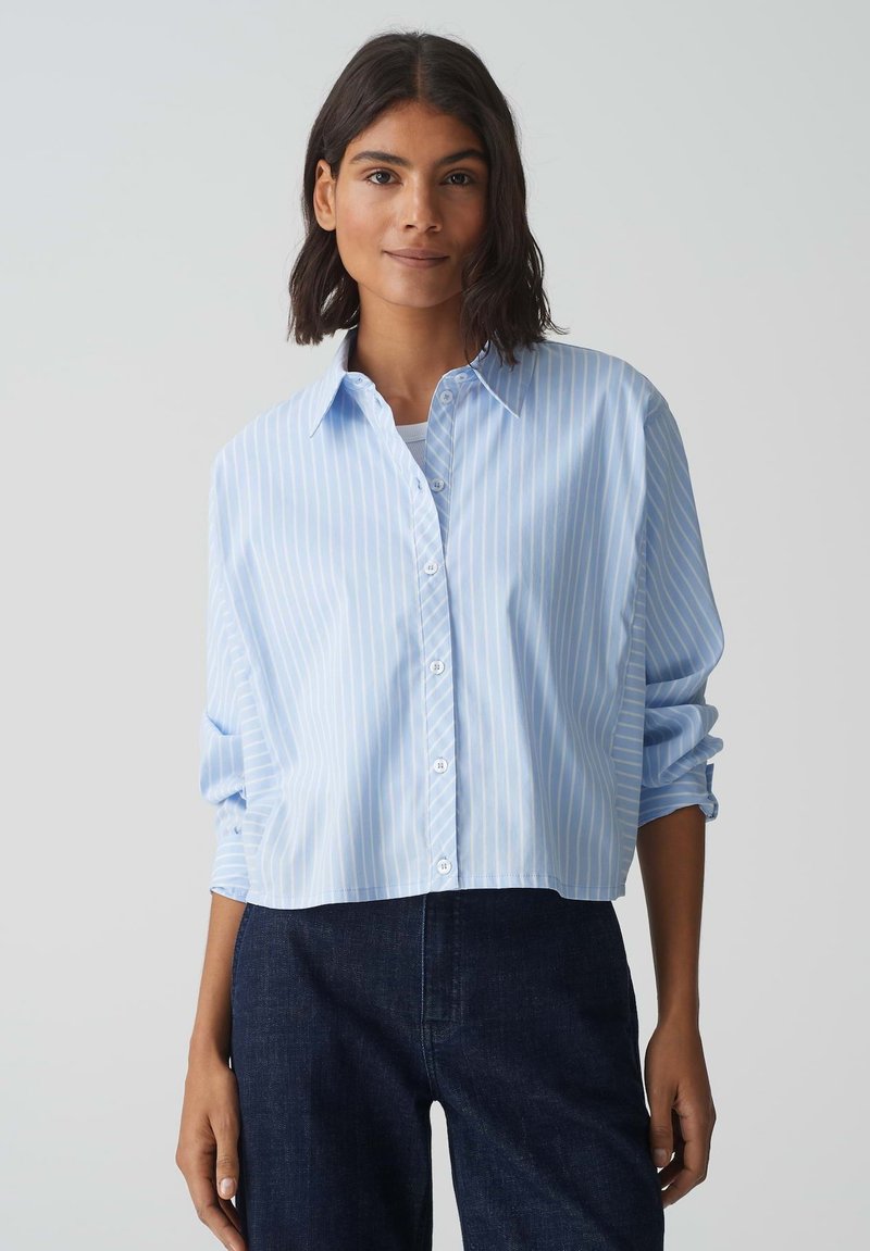 Camicia croppata blu chiaro con strisce verticali bianche, maniche lunghe arrotolate, realizzata in un tessuto liscio. Indossata con jeans scuri.
