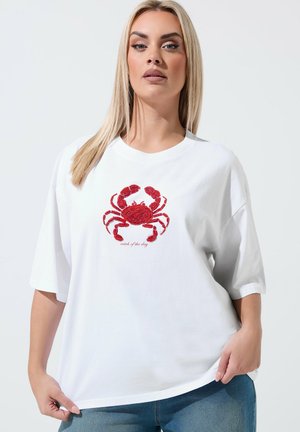 Blond kvinde iført hvid oversized T-shirt med rødt krabbedesign og teksten "fangst af dagen", kombineret med blå jeans mod ensartet baggrund.
