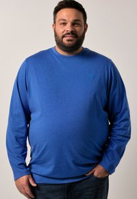 Muž se vztyčeným plnovousem, oblečený v modré košili s dlouhým rukávem a tmavých džínách, stojící s jednou rukou v kapse na jednobarevném pozadí.