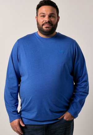 Hombre con barba vistiendo una camisa de manga larga azul y jeans oscuros, de pie con una mano en el bolsillo frente a un fondo liso.