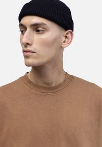 Brown crewneck sweatshirt made of soft cotton, featuring a ribbed collar. Worn with a dark, knitted beanie. Close-up on upper body and face.