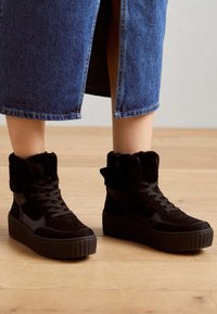 Baskets montantes noires avec un col en fausse fourrure texturée, une semelle plateforme et un laçage à l'avant. Matériaux lisses et en daim avec un design élégant.