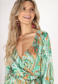 Rochie cu un model verde și portocalie, cu un decolteu adânc în V și un design înfășurat. Mâneci lungi, realizate dintr-un material moale și fluid. Cercei roz completează look-ul.