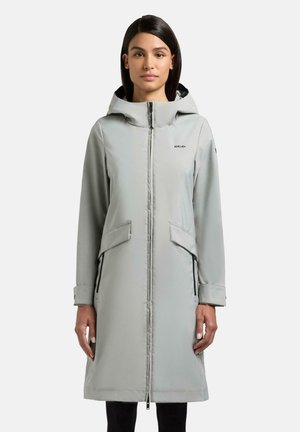 Kvinde iført en lysegrå lang hættetrøje med lynlås, stående med arme afslappet ned langs siden mod en ensfarvet hvid baggrund.